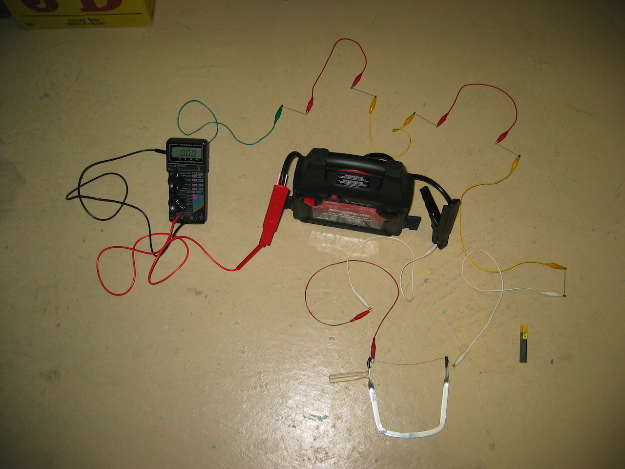 A series circuit of a 12 volt battery pack, 5 pencil leads, a multimeter, and a length of nichrome wire mounted in a small hacksaw where the saw blade should be. The elements of the circuit are connected with variously-coloured alligator clips.