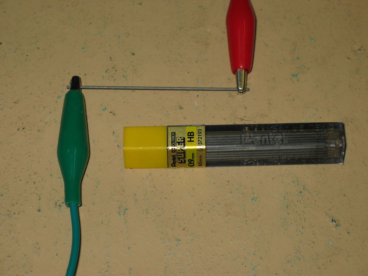 Close-up shot of a mechanical pencil lead with an alligator clip attached to each end, sitting adjacent to a tube of pencil leads.